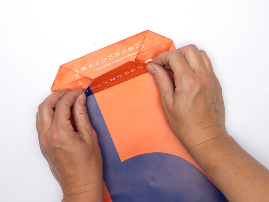 Tüte aus Papier falten ohne Vorlage: Jemand faltet den Boden einer Papiertüte aus einem rechteckigen, orangefarbenen Kalenderblatt von Athesia. Der Untergrund ist weiß. Foto: Johanna Rundel 