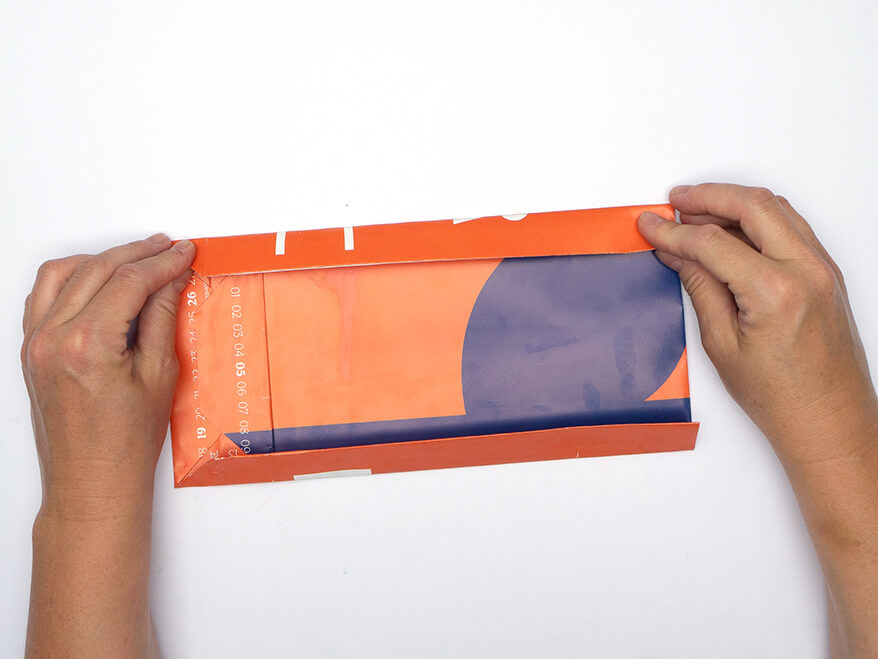 Tüte aus Papier falten ohne Vorlage: Jemand faltet die Seitenteile einer Papiertüte aus einem rechteckigen, orangefarbenen Kalenderblatt von Athesia. Der Untergrund ist weiß. Foto: Johanna Rundel 