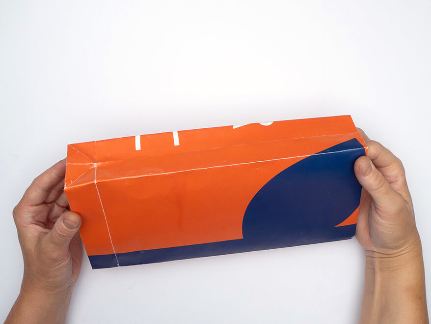 Tüte aus Papier falten ohne Vorlage: Jemand faltet eine, aus einem rechteckigen, orangefarbenen Kalenderblatt von Athesia gefalteten Papiertüte auseinander. Der Untergrund ist weiß. Foto: Johanna Rundel 