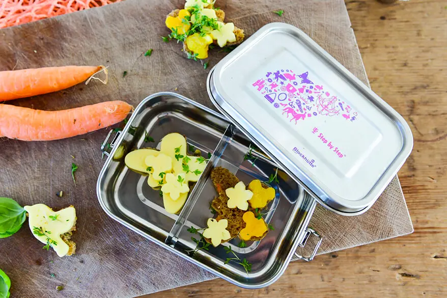 Eine geöffnete Brotdose aus Edelstahl von Step by Step liegt auf einem Holzbrett auf einem Holztisch. Sie ist geöffnet. Darin sind Käsebrote in Schmetterlingsform und Rohkost in Blütenform zu sehen. Neben der Brotdose liegen ein paar Karotten.
