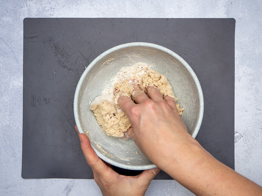 Jemand knetet in einer kleinen Rührschüssel, die auf einer dunkelgrauen Backmatte steht, Mehl, Wasser, Salz und Öl zu einem Salzteig zusammen. Der Untergrund ist Betongrau. Foto: Johanna Rundel 