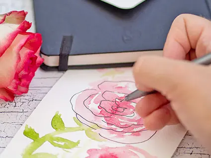 Jemand malt mit Aquarellfarben rote Rosen auf eine Aquarellpostkarte von Hahnemühle. Im Hintergrund liegt ein schwarzes Sketchbook, auf dem eine weitere Postkarte liegt.