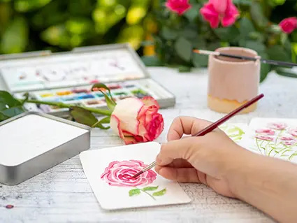 Jemand aquarelliert in der Natur eine Rose auf eine Aquarellpostkarte von Hahnemühle. 