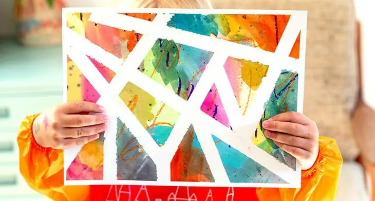 Ein Kleinkind in einer orange-roten Malerschürze von Faber-Castell hält ein Tape-Resist-Art-Kunstwerk in die Kamera. Foto: Johanna Rundel