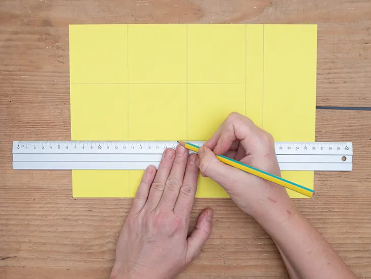 Jemand zeichnet mit einem Lineal und Bleistift Linien auf einen gelben Fotokarton von HEYDA. Foto: Johanna Rundel)