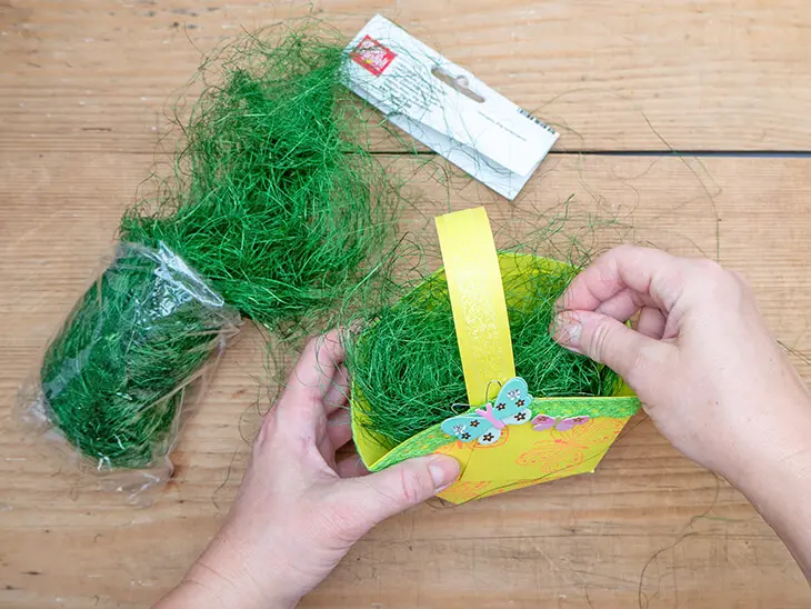 Jemand befüllt ein selbstgebasteltes, Osternest aus gelbem Papier von HEYDA mit grünem Ostergras von KNORR prandell. Foto: Johanna Rundel
