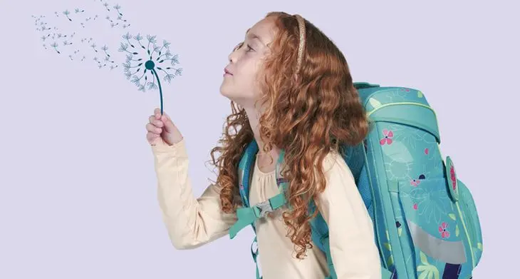 Ein Mädchen mit einem Schulranzen von DerDieDas auf dem Rücken „pustet“ eine gezeichnete Pusteblume an