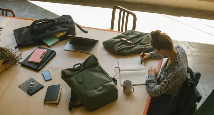 Blick von oben auf einen Schreibtisch, an dem eine Frau in ein Notizbuch von Moleskine schreibt, um sie herum ist Reisegepäck verteilt.