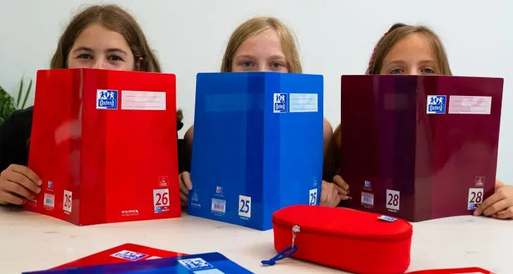 Drei Schulkinder verstecken ihre Gesichter zur Hälfte hinter Schulheften von Oxford