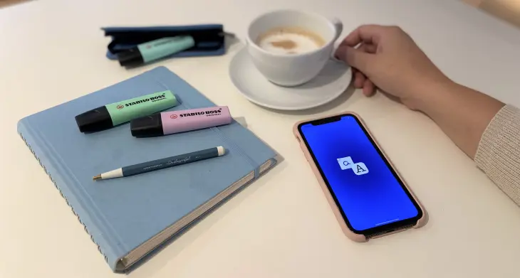 Auf einem Tisch liegt ein zugeschlagenes Notizbuch. Darauf liegen 2 Textmarker der Marke Stabilo und ein LEUCHTTURM1917 Drehgriffel. Daneben liegt ein Smartphone und jemand greift nach einer Tasse, die daneben steht.