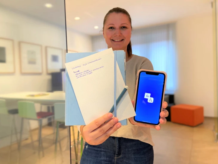 Eine junge Frau hält lächelnd ein Notizbuch mit Stift und ein Smartphone, auf dem die App Pen to Print läuft.