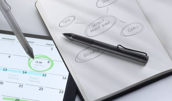 Auf einem Tisch liegt ein aufgeklapptes Notizbuch, in dem eine Mindmap skizziert ist, darauf liegt ein Lamy Safari Twin Pen All Black EMR. Neben dem Notizbuch liegt ein eingeschaltetes Tablet, auf dem ein Kalender zu sehen ist. Ein Termin wurde vion einem Lamy Safari Twin Pen All Black EMR grün umkreist.