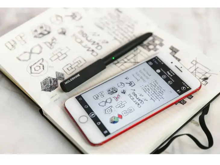 Ein Moleskinebuch liegt aufgeklappt auf einem Tisch. Darauf liegen ein Smartphone und ein Smart Pen. MIt Hilfe dieses Moleskine Smart Writing Systems kann man Handschrift digitalisieren.