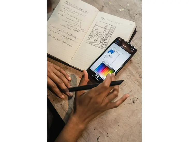 Auf einem Tisch liegt ein aufgeklapptes Notizbuch, daneben ein Smartphone. Auf dem Smartphone sieht man exakt die gleiche Seite des Notizbuchs, die mit Hilfe des Smar Writing Systems von Moleskine digitalisiert wurde. Eine Hand hält den Smart Wrtiting Pen und tippt gleichzeitig auf das Display.