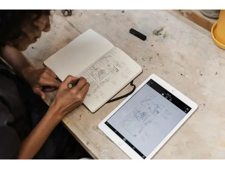 Eine Person hält den Moleskine Smart Pen in der Hand und malt damit Skizzen in ein Moleskine Notizbuch. Daneben liegt ein Tablet, das die Skizzen sofort digital darstellt.