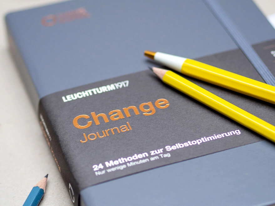 Auf einem grauen Untergrund liegt das blaue Change Journal von Leuchtturm1917. Auf dem Journal liegen ein gelber Drehgriffel und ein gelber Bleistift. Weitere blaue Bleistifte sind um das Journal verteilt. Foto: Johanna Rundel