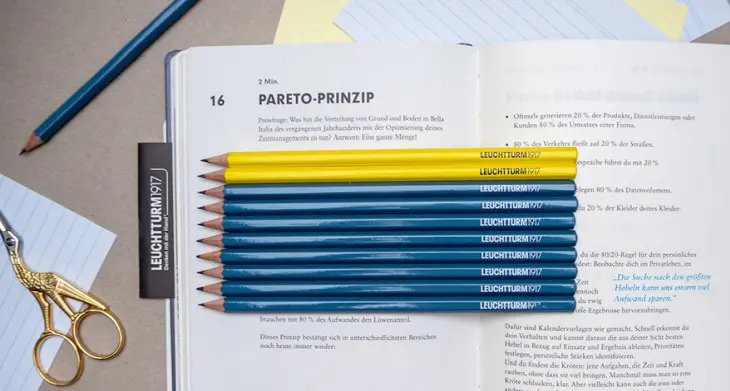 Das Pareto-Prinzip wird in einem blauen Change Journal von Leuchtturm1917 erklärt. Die Seiten über das Paretoprinzip sind aufgeschlagen. Auf dem Journal liegen zwei gelbe und acht blaue Bleistifte. Um das Journal sind weitere Büromaterialien verteilt. Foto: Johanna Rundel