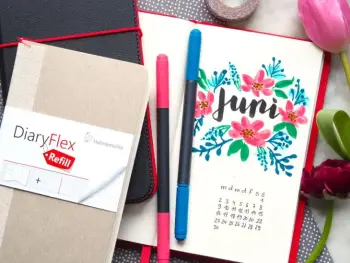 In ein aufgeschlagenes Notizbuch von Hahnemühle ist das Wort „Juni“ gelettert, umgeben von gemalten Blumen.