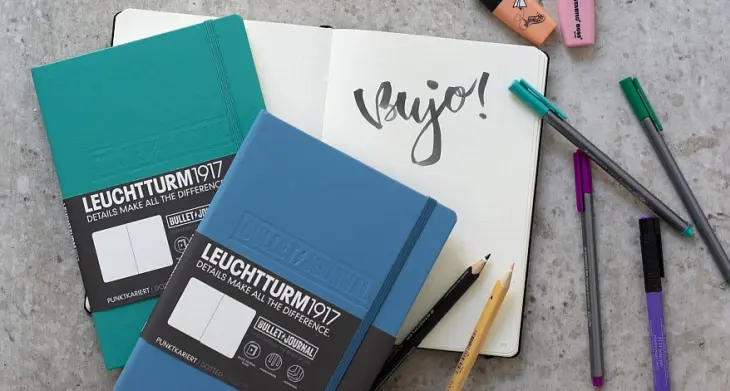 Auf einem Tisch liegen Bullet Journals von LEUCHTURM1917 umgeben von Stiften, auf eine aufgeklappte Seite ist das Wort BuJo in Lettering geschrieben. 