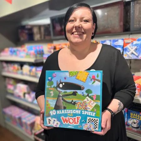 Eine Mitarbeiterin von Ortloff hält eine Spielesammlung in der Hand und lächelt in die Kamera.