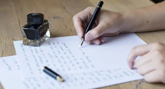 Eine Hand schreibt mit einem Füllfederhalter einen Brief, auf dem Papier steht ein Glas Tinte.