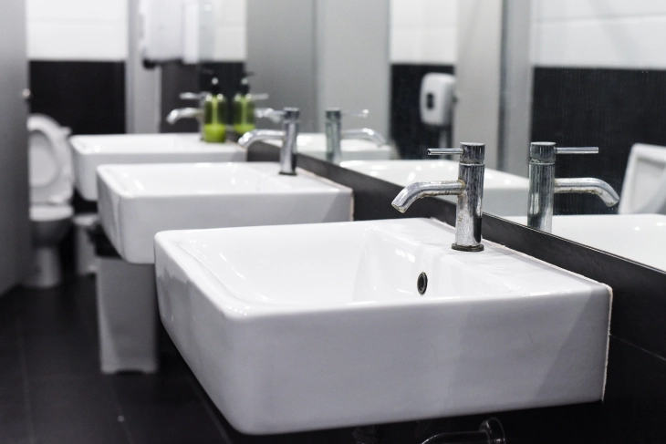 Das Bild zeigt einen modernen, geschmackvollen Waschraum mit drei weißen Waschbecken. Im Hintergrund befinden sich Toilettenkabinen mit Toilettenpapierspendern.