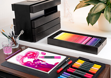 Auf einem Tisch Stifteetuis der Art&Graphic-Collection von Faber-Castell. Pinkes, abstraktes Bild, mit Aquarellstiften gemalt.