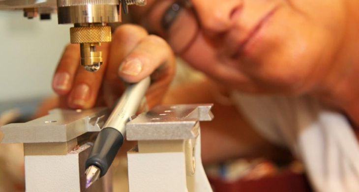 Ein Füller liegt in einer Halterung einer Vorrichtung zum Gravieren. Im Hintergrund ist unscharf eine frau zu erkennen, die in die Kamera lächelt.