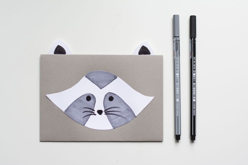 Bastelanleitung Einladungskarte zum Kindergeburtstag: Selbst gebastelte Do it yourself-Karte als Waschbär, Schritt 3: Mit Filzstift wird graues Gesichtsfell, Augen, Schnauze und Barthaare aufgemalt.