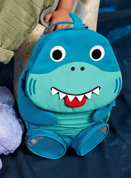 Ein Kindergartenrucksack in Hai-Form von Affenzahn steht neben einem Stuhl, auf dem ein Kind sitzt.