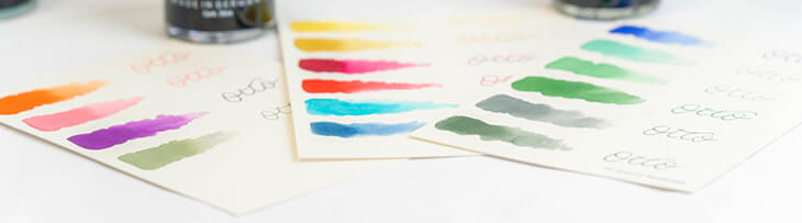 Auf einem hellen Untergrund stehen verschiedenfarbige Tintenfässer von Otto Hutt. Drei Musterkarten mit Farbstrichen liegen dazwischen.