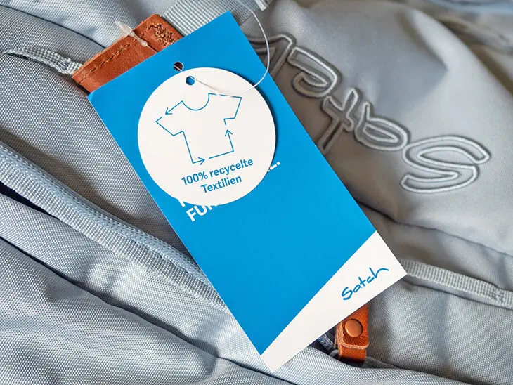 Ein Tag auf dem ein T-Shirt zu sehen ist und auf dem 100% recycelte Textilien steht, ist an einem blauen satch Schulrucksack befestigt. Foto: Johanna Rundel