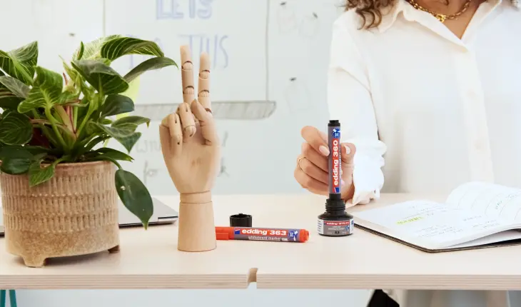 Eine Person füllt einen edding Marker nach. Die Nachfüllflasche steht auf einem hellen Schreibtisch mit einer Grünpflanze, einem Laptop einer Hand aus Holz, einem roten edding Marker und einem Notizbuch.