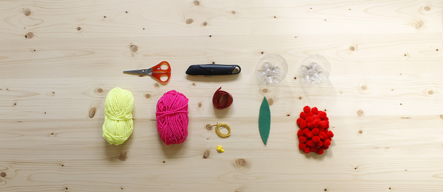 Foto aus der Vogelperspektive auf einen Holztisch, auf dem eine Schere, ein Cuttermesser, zwei untere Hälften von Plastikflaschen, ein glebes und ein pinkes Wollknäuel, Schleifenband, grüner Filz und rote Bommel liegen. 
