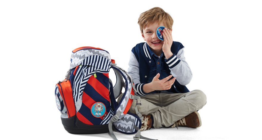 EinJunge im Grundschulalter sitzt auf dem Boden. Neben ihm steht ein ergobag Schulranzen. Der Junge hält sich ein ergobag-Klettie vor ein Auge und lacht.