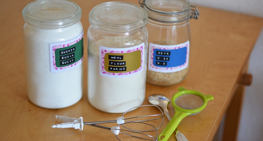 Beschriftete Vorratsgläser mit Zucker, Mehl und Reis auf einem Holztisch.