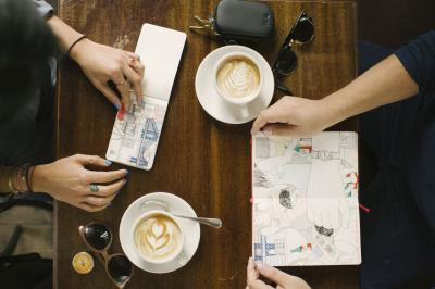 Foto von oben auf einen Tisch, an dem zwei Frauen bei einem Kaffee vor ihren aufgeschlagenen und beschriebenen und gestalteten Notizbüchern sitzen.