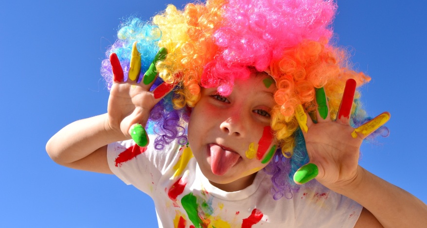 Ein Kind im Grundschulalter trägt eine bunte Clown-Perücke, seine Hände, das Gesicht und das T-Shirt sind mit grüner, roter und gelber Farbe bemalt. Das Kind hält sie Hände neben das Gesicht und streckt dir Zunge aus dem Mund.