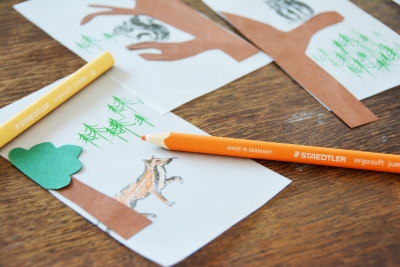 Auf einem Tisch liegen 3 gebastelte und gemalte Walöd-Motive mit einem Baum und einem Fuchs. Darauf liegen ein gelber und ein oranger Buntstift.