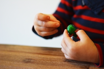 Ein Kleinkind hält in der linken Hand eine grüne runde Kugel, die zu einem Mäusekörper geformt ist und braune Ohren hat. In der rechten Hand hält das Kind einen bruanen Schwanz aus Knetbienenwachs.