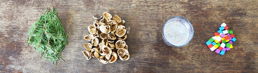 Foto aus der Vogelperspektive auf einen Holztisch, auf dem in kleinen Portionen Tannennadeln, Nuss-Schalen, Sand und kleine bunte Holzklötze liegen.