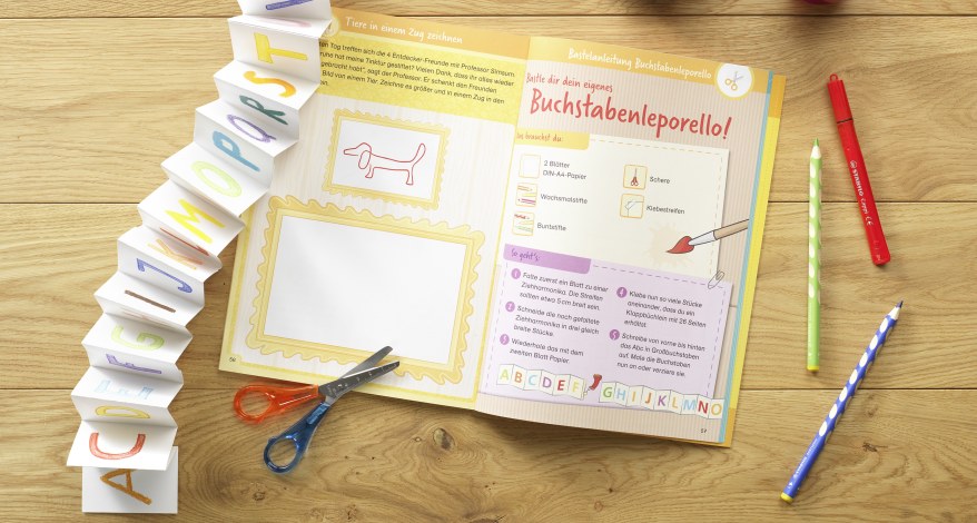 Ein Schreibübungsheft liegt aufgeschlagen auf einem Tisch, daneben Buntstifte und eine Buchstaben-Girlande.