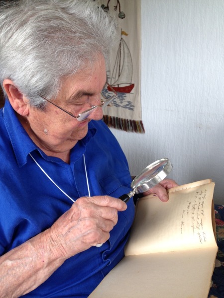 Eine ältere Dame mit grauen Haaren und Brlle hält eine Lupe in der Hand. Mit dieser inspiziert sie ein altes Heft, in welches mit Kurrentschrift geschrieben wurde.