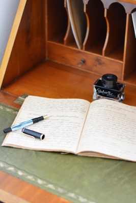 Auf einem alten Sekretär aus Holz liegt ein vollgeschriebenes Heft. Darauf ist ein offener Füller der Marke Pelikan zu sehen. Über dem Heft steht ein Tintenglas mit schwarzer Tinte der Edelstein Ink Collection von Pleikan.