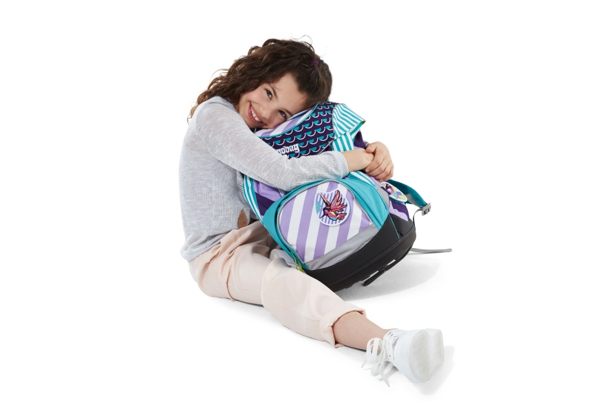 Mädchen sitzt am Boden und umarmt freudig ihren bunten ergobag-Schulrucksack.