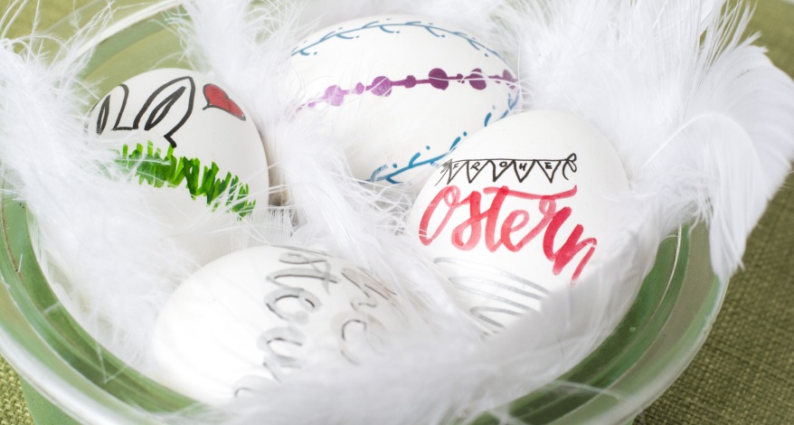 Ostereier mit Schrift und Zeichnungen in Schale mit weißen Federn