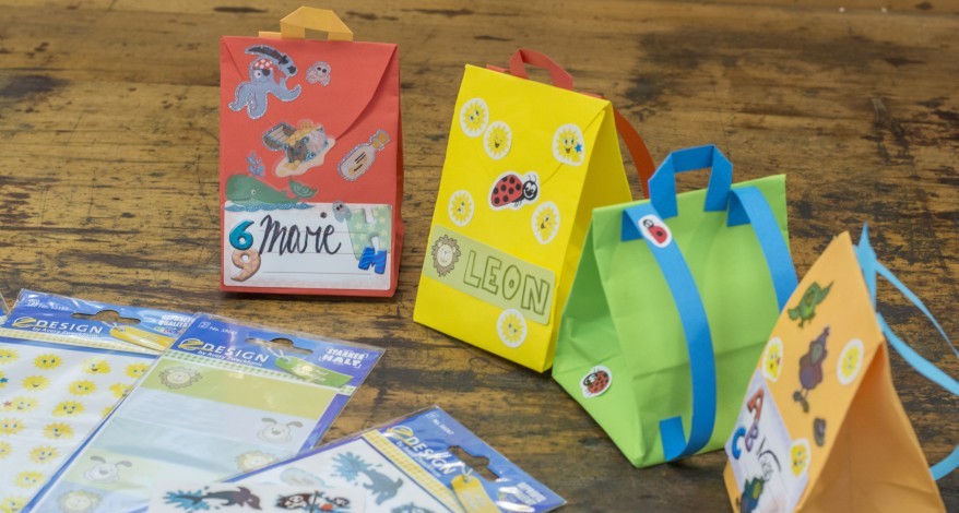 Vier bunte Geschenktüten in Form eines Schulranzens stehen auf einem alten Holzboden. Die Tüten wurden mit Stickern und Etiketten beklebt und mit Namen beschriftet. In der linken unteren Bildecke liegen einige gefüllte Stickerverpackungen von Avery Zweckform. 