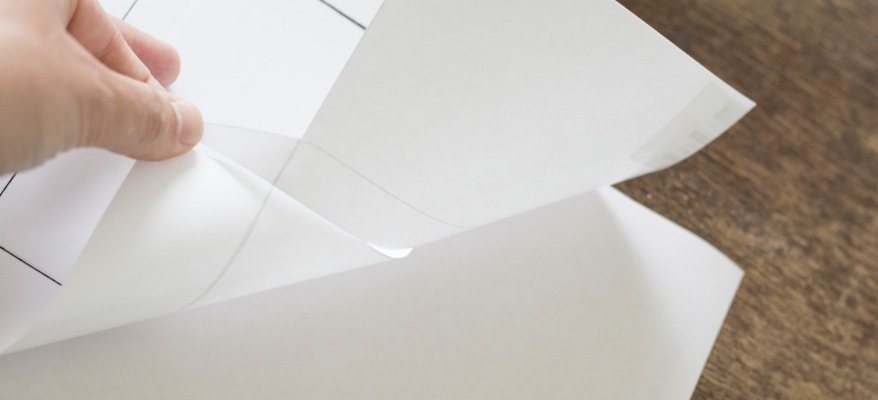 Eine Hand zieht eine Schutzfolie von selbstklebenden Papier, das auf einem Holztisch liegt.