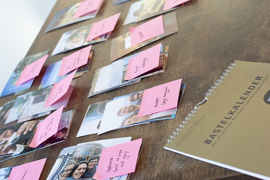Auf einem Holztisch liegen verschiedene Fotostapel, die mit Haftnotizen beschritet wurden. Im rechten Bildrand liegt ein Bastelkalender von Heye.