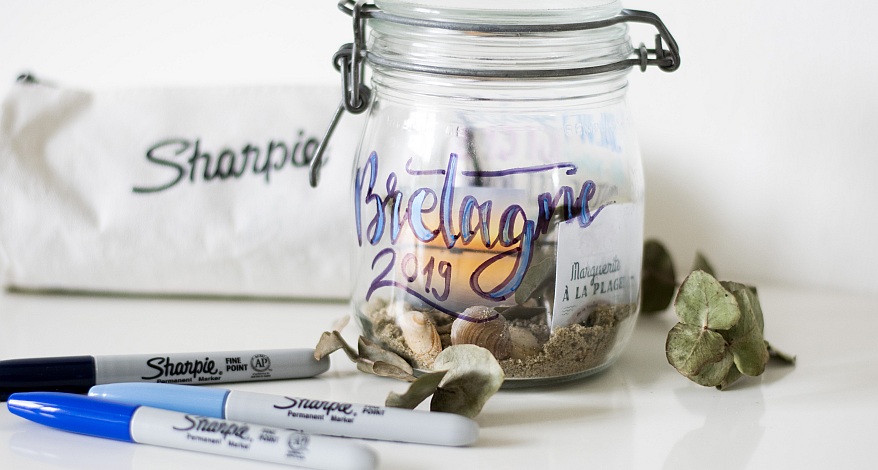 Auf einem Tisch steht ein Weckglas, welches in blau mit der Aufschrift "Bretagne 2019" belettert ist. Das Glas ist mit Steinen, Sand und Muscheln befüllt. Vor dem Glas liegen der Marker der Marke Sharpie in schwar, hellblau und dunkelblau. Im Hintergrund ist ein Schlampermäppchen mit der Aufschrift "Sharpie" zu sehen.
